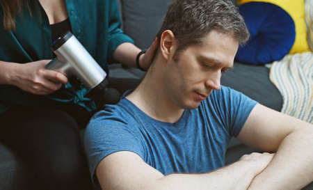 Woman massaging man with massage percussion device at home.の写真素材