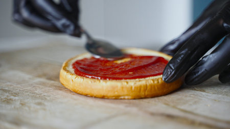Chef in black gloves smears a bun with ketchup for hamburger.