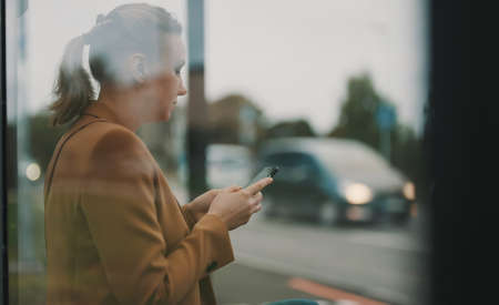 Woman with smartphone is waiting for bus at bus stop.の写真素材