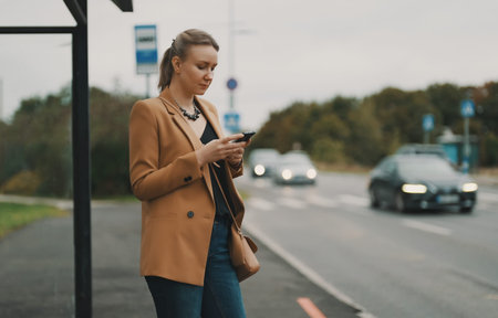 Woman with smartphone is waiting for bus at bus stop.