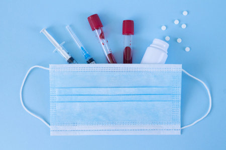 Blood test tubes, antiviral medical mask, syringe and pills on blue background top viewの写真素材