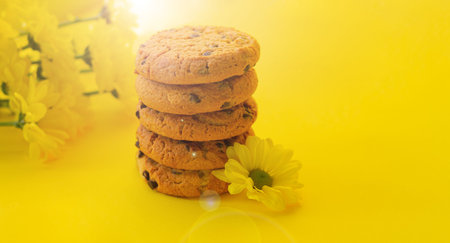 Stack of delicious chocolate chip cookies next to flowers on yellow background