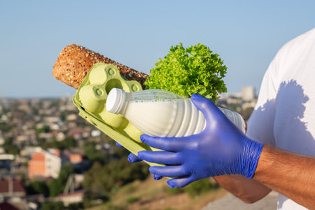 Male courier in protective gloves holding fresh food on city landscape background. Delivery during pandemicの写真素材