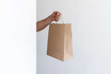 Close-up man holds in hand brown clear empty blank craft paper bag for takeaway on light gray background. Packaging and delivery service conceptの写真素材
