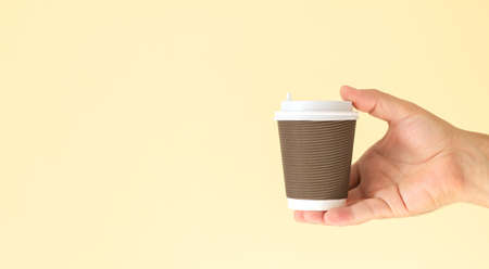 Man's hand holding brown paper coffee cup with white plastic cap isolated on beige background with copy spaceの写真素材
