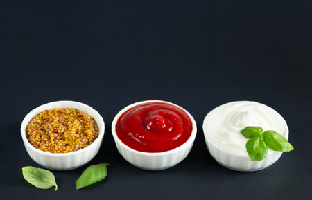 Set of popular sauces: ketchup, dijon mustard and mayonnaise in small white bowls on black...