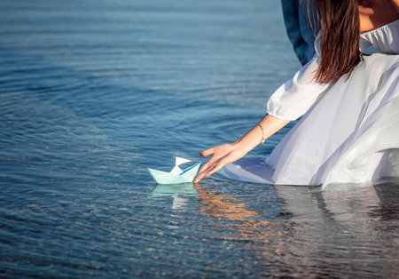 Young woman in white dress holds light blue paper boat in hand on sea backgroundの写真素材