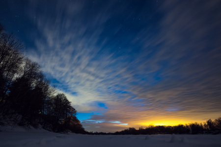 Snow-covered river near the forest on a winter frosty night with clouds and stars in the sky.And a trace from the planeの写真素材