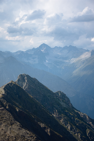 The peaks of the Caucasus Mountains in cloudy clouds and the rays of the sun through them in the outskirts of Dombai. And on the tops of snow lies.の写真素材
