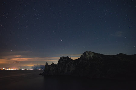 A large panorama of a beautiful view of the sea among the mountains and a beautiful starry sky with the Milky Way. The city by the sea glows at night in the distanceの写真素材