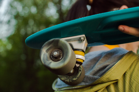 Girl in green outwear carrying blue plastic penny board on her shoulder. Side view. The concept of modern culture, penny skateboarding, outdoor activity, modern lifestyle.の写真素材