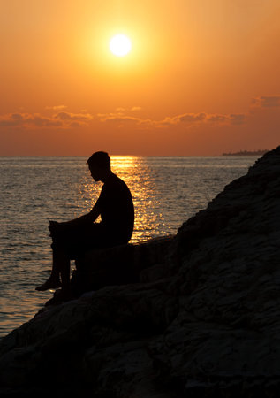 Silhouette of man from side sitting on a rock and enjoying the sunsetの写真素材