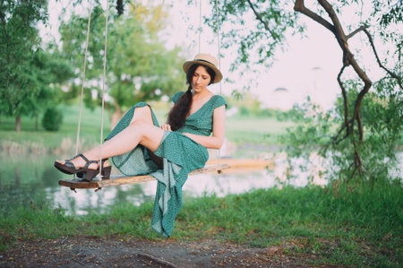 beautiful girl in a green dress and hat is resting on a swing against the backdrop of the lake