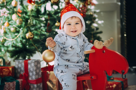 little baby boy in a Christmas cap sitting on a wooden horse by the Christmas tree