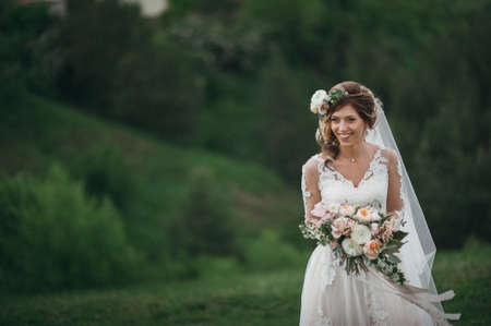 Beautiful bride with fresh flowers in her hair and a bouquet posing on a green fieldの写真素材