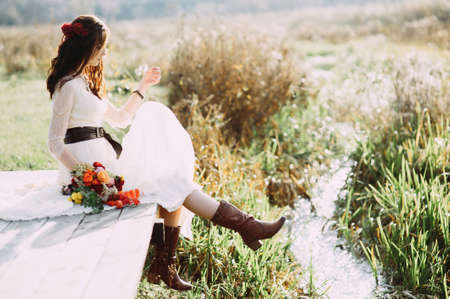 beautiful bride with a bouquet and in boots sits on a bridge against the background of the lakeの写真素材