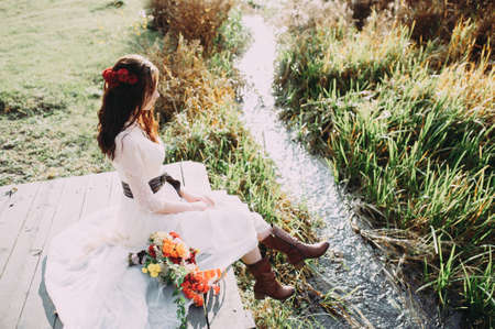 beautiful bride with a bouquet and in boots sits on a bridge against the background of the lakeの写真素材