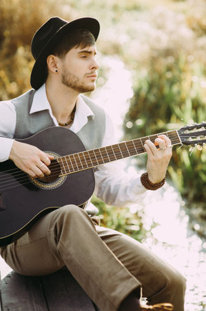 Young man playing guitar sitting on the bank of a mountain river on a background of rocks and...