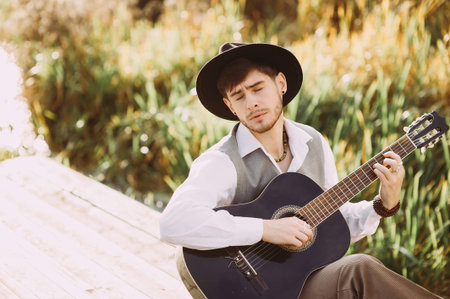 Young man playing guitar sitting on the bank of a mountain river on a background of rocks and...