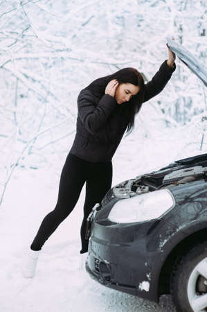 beautiful girl near the motor of a broken car on a winter snowy road. calls for helpの写真素材