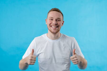 The guy smiles at the camera and shows like. Guy in a white T-shirt on a blue background. Portrait of a guy of European appearance. Fashionable man.の写真素材