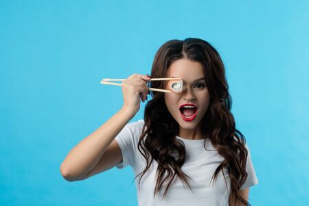 Portrait of a brunette girl who has hadi sushi in her hands. The girl opened her mouth wide in surprise. She brought sushi to her eyeの写真素材