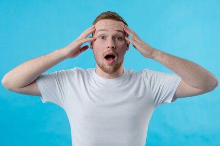 The red-haired guy holds his head in surprise. Red-haired guy in a white T-shirt with open mouth. Fashion, beauty, model, studio conceptの写真素材