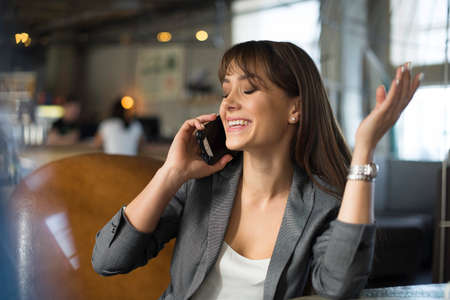 Young happy woman talking on mobile phone with friend while sitting alone in modern coffee shop interior, smiling hipster girl calling with cell telephone while relaxing after walking in summer dayの写真素材