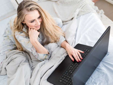 Blonde woman works on her laptop lying on the bed in the morning