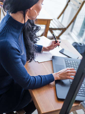 Young business woman in gray dress sitting at table in cafe and writing in notebook. On table is laptop, smartphone and cup of coffee. Freelancer working in coffee shop. Student learning onlineの写真素材