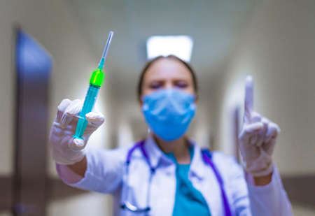 Young female doctor holding a syringe in the hospital corridor and showing attention sign. Selective focus on the injectorの写真素材