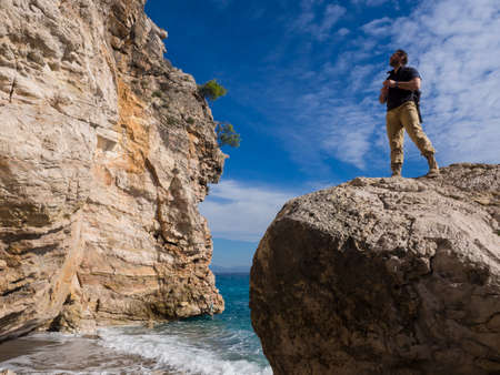 Traveler with backpack on the rocks near the sea looking away. Summer Travel Vacation. Handsome young caucasian tourist man in casual clothes outdoors on the nature.の写真素材