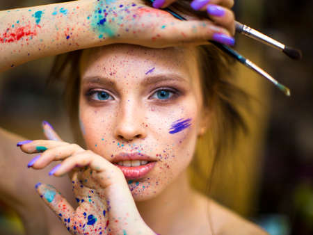 Close up portrait of beautiful female artist with dirty hands with different paints on them, holding paint brushes near her face and eyes.の写真素材