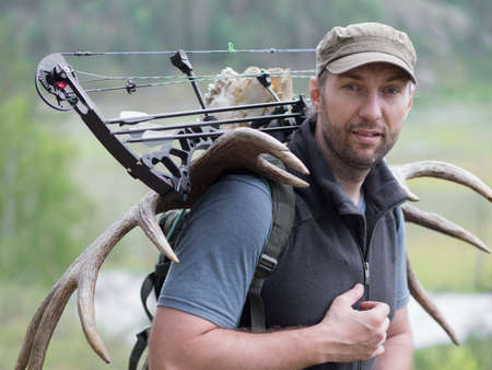 A hunter with a bow in the woods carries moose horns on his back and looking at camera.