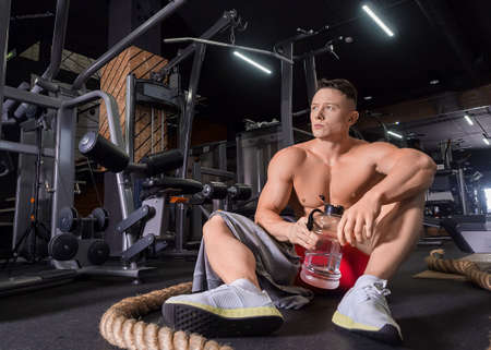 Tired man sits on a floor after hardworking in gym drinking water.