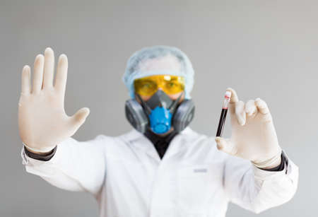 Photo of a doctor in protective clothing holds a test tube with a sample of infected blood in his hand and showing stop sign by another hand.の写真素材