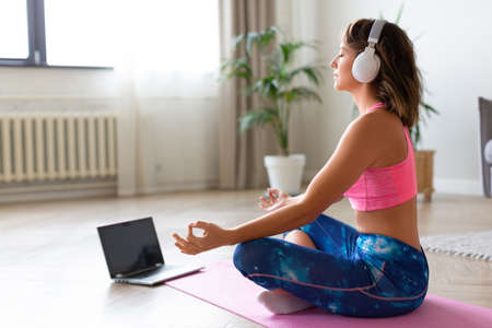 Online yoga lesson. Woman in headphones in lotus position in front of laptop monitor.の写真素材