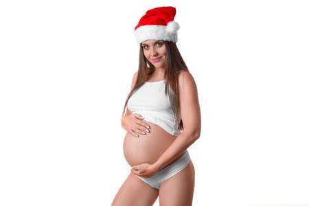 Smiley pregnant woman in Santa hat gently holds her belly over white background.の写真素材