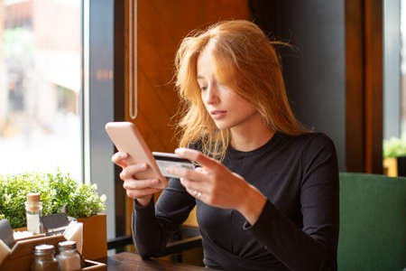 Red haired young woman making card payment through mobile phone to pay bills.