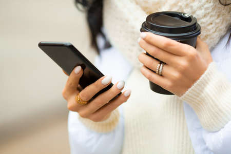 Female hands with smartphone with a mug of coffee outdoor.
