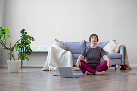 Photo of a senior woman meditaing in front of laptop monitor. Online yoga lesson.の写真素材
