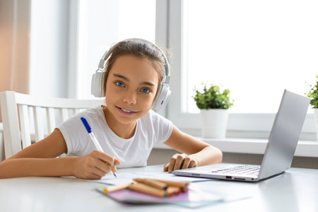 Teenager girl doing homework at home in front of a laptop monitor. She is looking at the camera and smiling.の写真素材