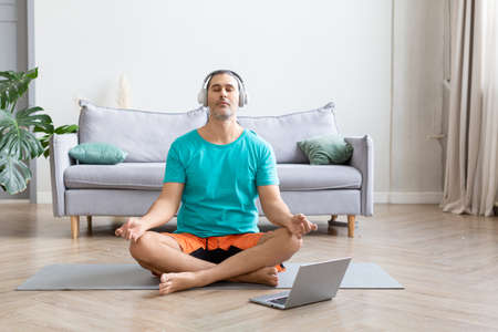 Middle-aged man is meditating at home in front of his laptop - he is listening to relaxing music from an online yoga tutorial.の写真素材