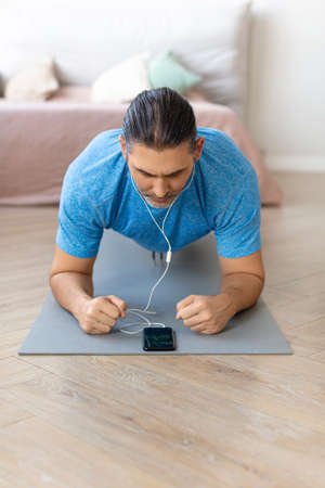 A man performs a plank exercise at home. He uses a fitness app on his smartphone.