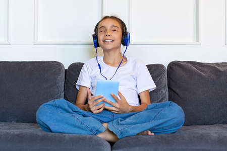 A teenage girl sits on the couch with a tablet and listens to music through headphones.