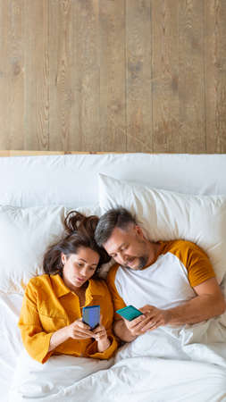 Vertical photo of a middle aged couple lying in bed with smartphones.の写真素材