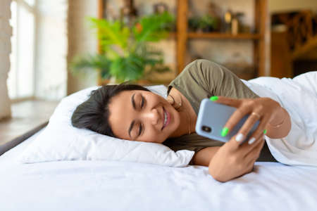 An asian woman lies in bed with cell phone. She woke up and was checking social media posts.の写真素材