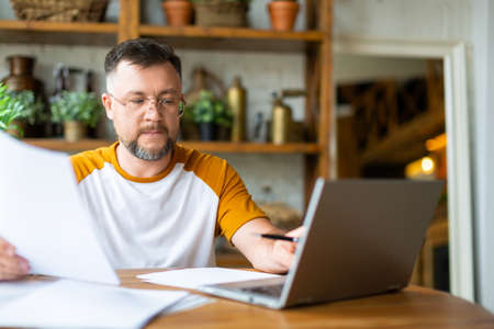 A middle-aged man works with papers at home in front of a laptop monitor. He is sitting with glasses...