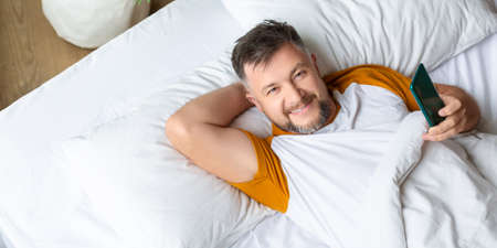 Smiling middle aged man with smartphone looking at camera. He lies in bed with a smartphone and...