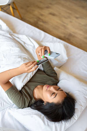 Asian woman doing online shopping while lying in bed. She holds a smartphone in her hands and credits the card and dials the card number in the online store. Vertical photo.の写真素材